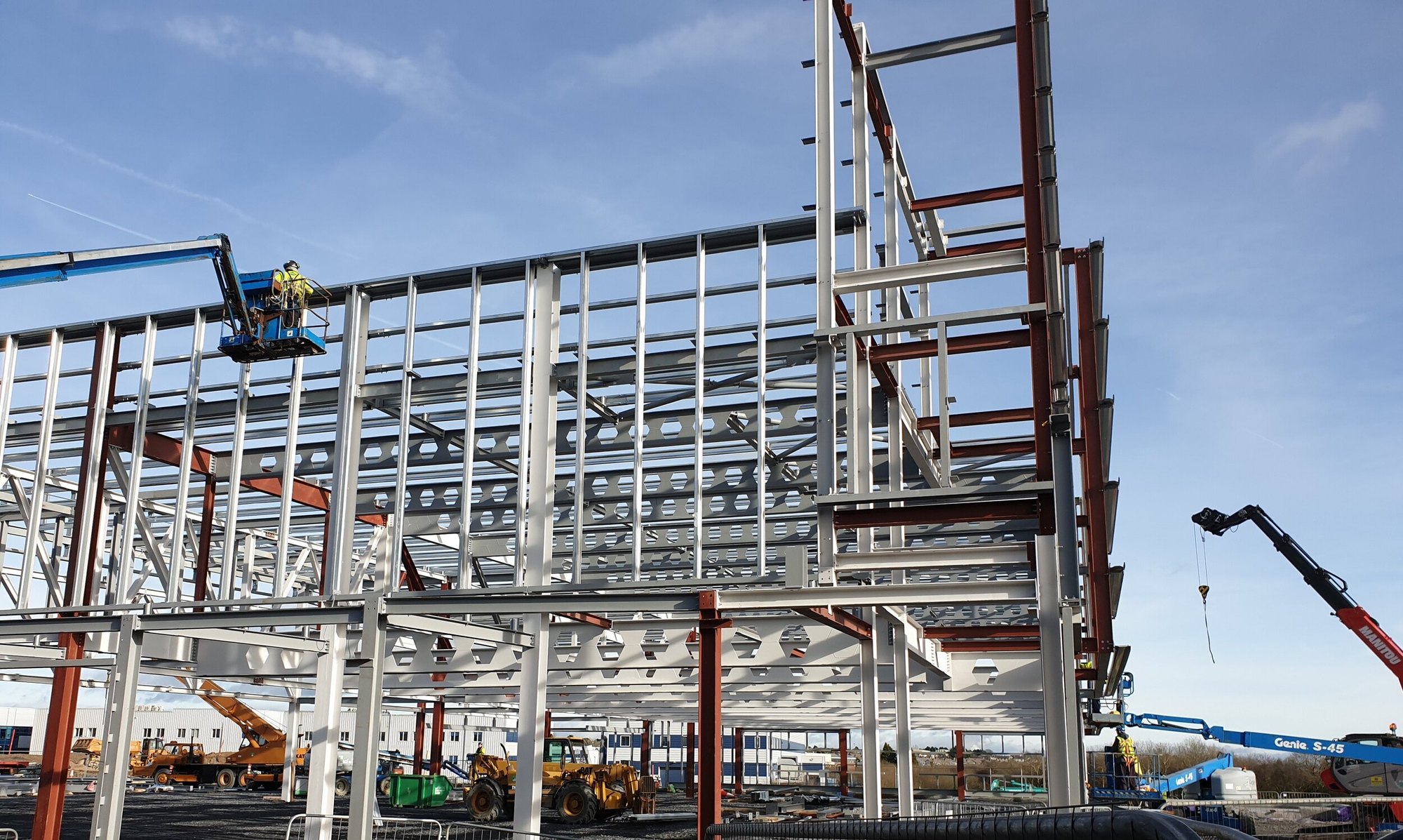 Structural steel frame going up with a cherry picker and a mobile crane on site under a blue sky